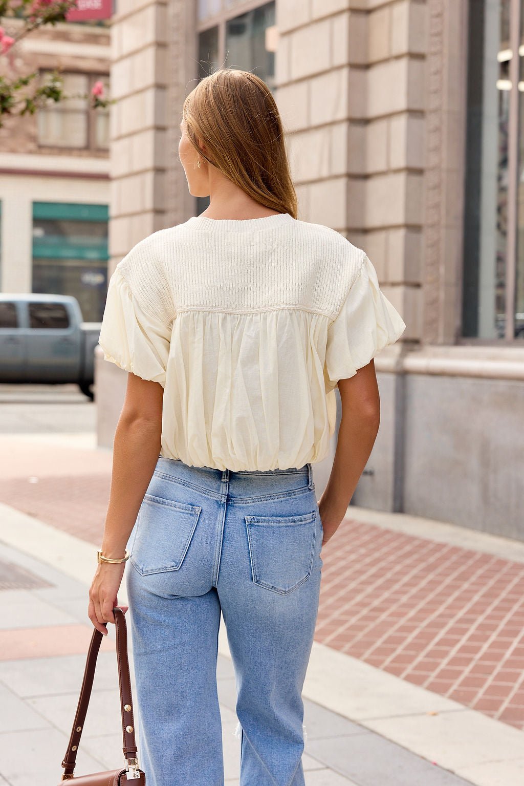 Woman with long hair in light jeans and Bubble Hem Short Sleeve Top - Ivory stands on a city sidewalk, holding a brown handbag.