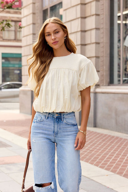 Woman with long wavy hair wears an ivory Bubble Hem Short Sleeve Top and ripped jeans, standing on a city sidewalk.
