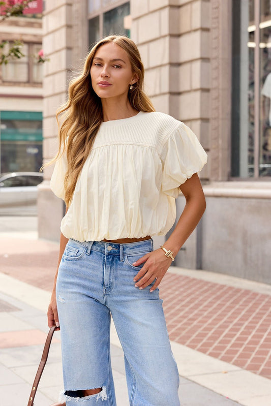 Woman with long hair wears the Bubble Hem Short Sleeve Top in Ivory and ripped jeans, holding a brown bag on the city sidewalk.
