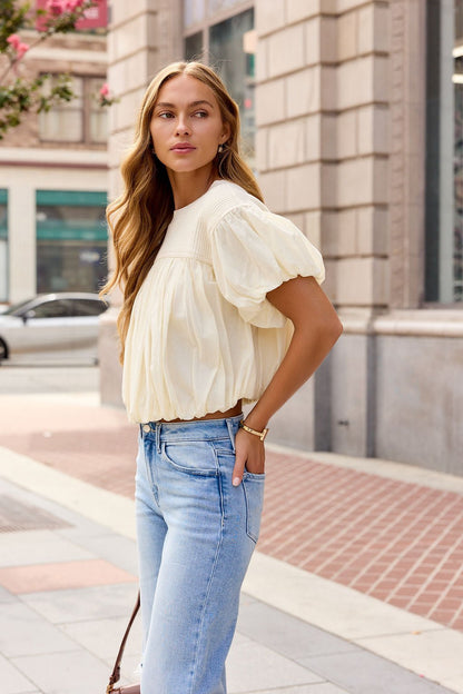 Woman with long wavy hair wears an ivory Bubble Hem Short Sleeve Top and light blue jeans, standing on a city sidewalk.