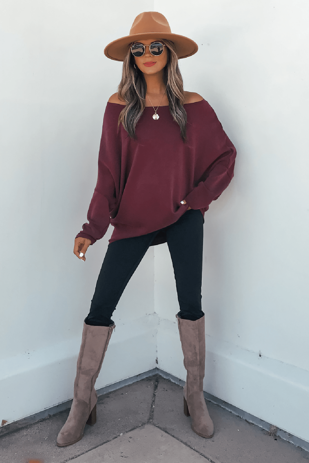 Woman in a tan hat, sunglasses, and Burgundy Soft Cloud Scuba Pullover poses by a white wall in black pants and taupe boots.