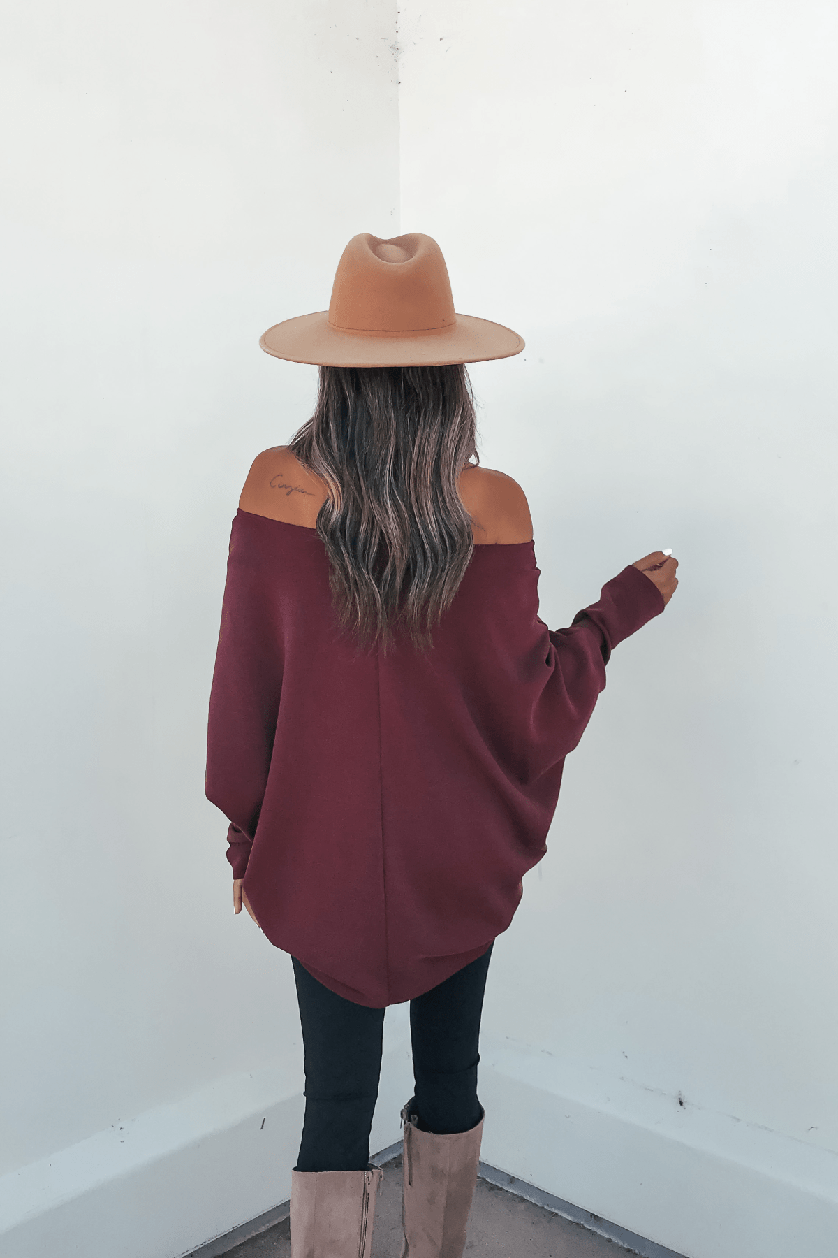 Woman in a tan hat and Burgundy Soft Cloud Scuba Pullover faces a white wall, showing her long wavy hair and tan knee-high boots.