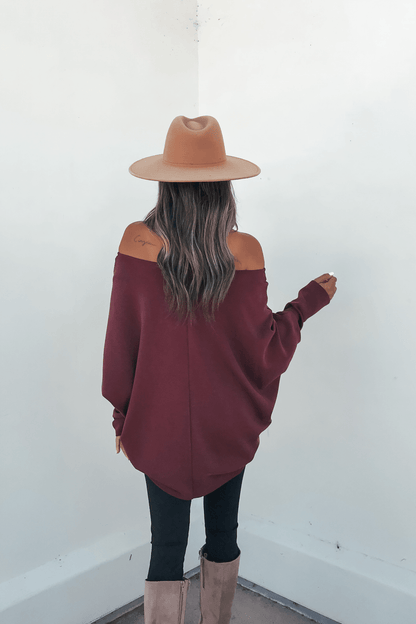 Woman in a tan hat and Burgundy Soft Cloud Scuba Pullover faces a white wall, showing her long wavy hair and tan knee-high boots.