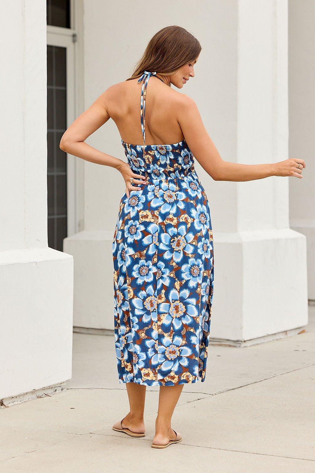 Woman wears Catalina Halter Midi Dress - Navy, featuring a smocked bust and floral print, standing outside with her back turned.