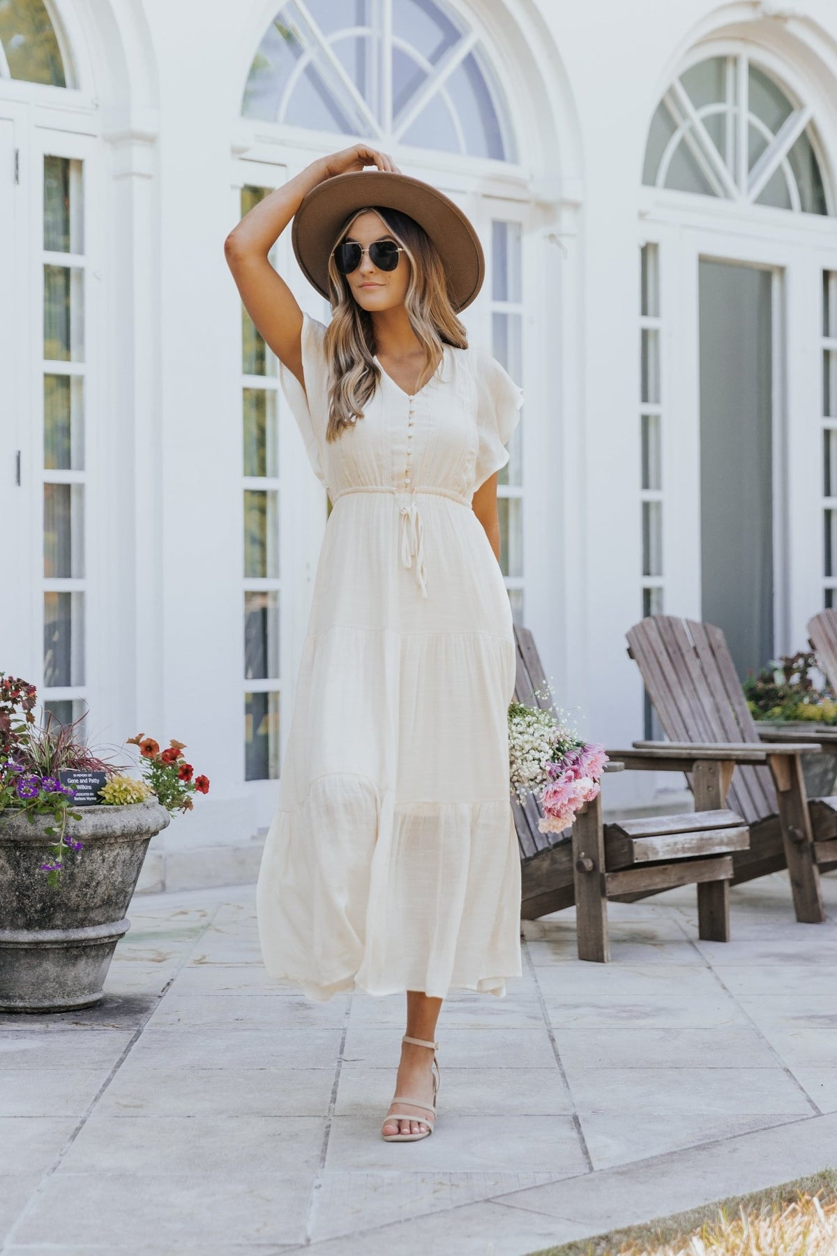Woman in the Celine Crochet Lace Tiered Boho Midi Dress walks past wooden chairs and flowers by a white building with arched windows.