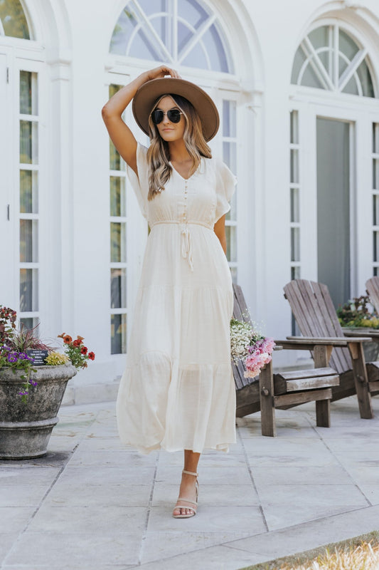 Woman in the Celine Crochet Lace Tiered Boho Midi Dress walks past wooden chairs and flowers by a white building with arched windows.