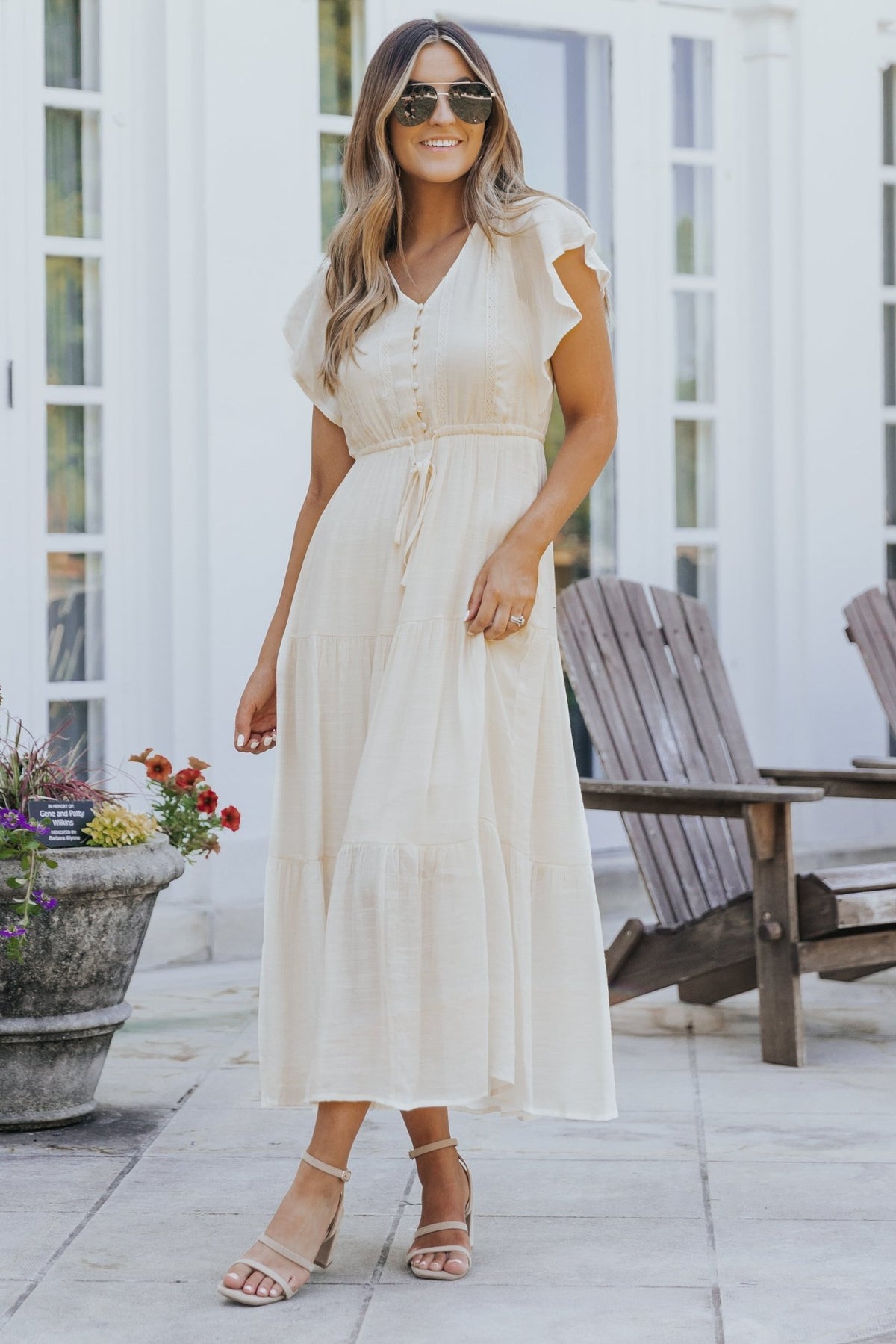 Woman in the Celine Crochet Lace Tiered Boho Midi Dress stands outdoors by wooden chairs and potted flowers, wearing sandals.