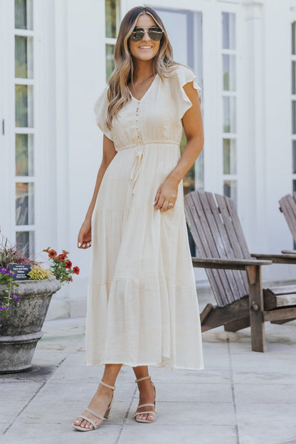 Woman in the Celine Crochet Lace Tiered Boho Midi Dress stands outdoors by wooden chairs and potted flowers, wearing sandals.