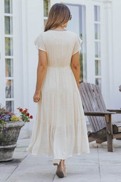 Woman in the Celine Crochet Lace Tiered Boho Midi Dress stands on a patio near wooden chairs and flowers, facing away.