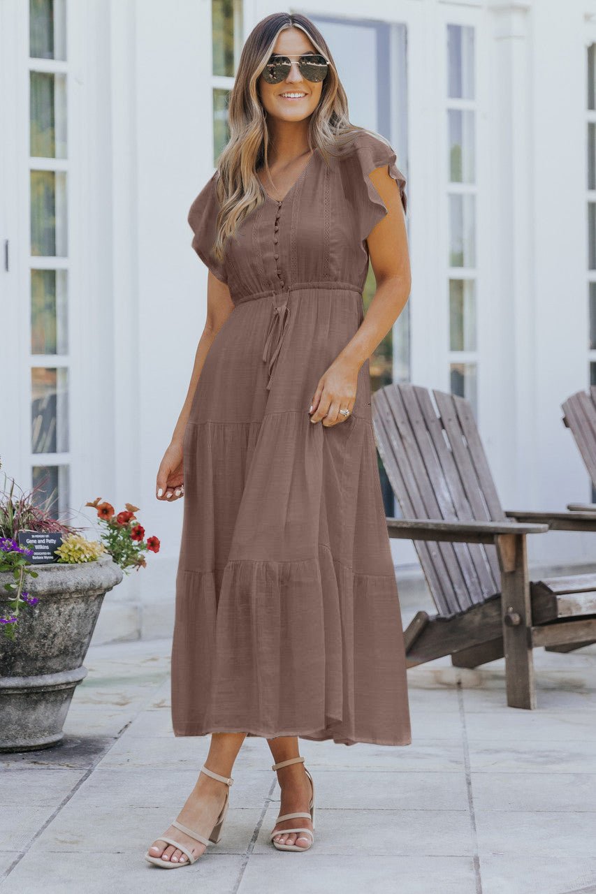 Woman in sunglasses smiles outdoors in the Celine Crochet Lace Tiered Boho Midi Dress - Brown, near flowers and wooden chairs.