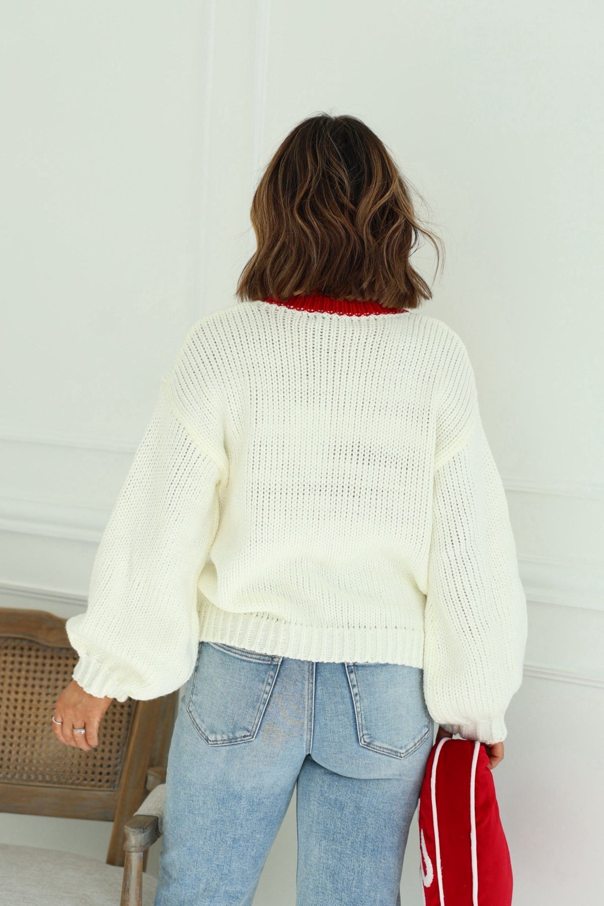 A woman with wavy brown hair stands with her back to the camera in a chunky cream Heart Shape Sweater and blue jeans.