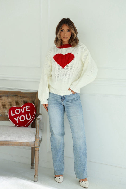 Woman in a Chunky Cream Heart Shape Sweater, blue jeans, and white shoes stands by a bench with a “LOVE YOU” pillow.