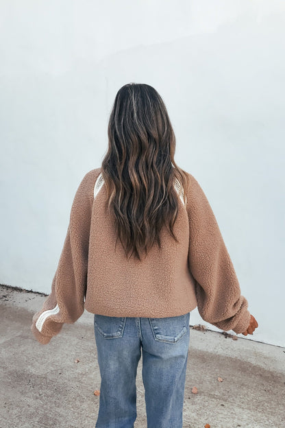 Person with wavy brown hair in a Contrast Quarter Zip Jacket - Sand and jeans, back to camera, against a light wall.