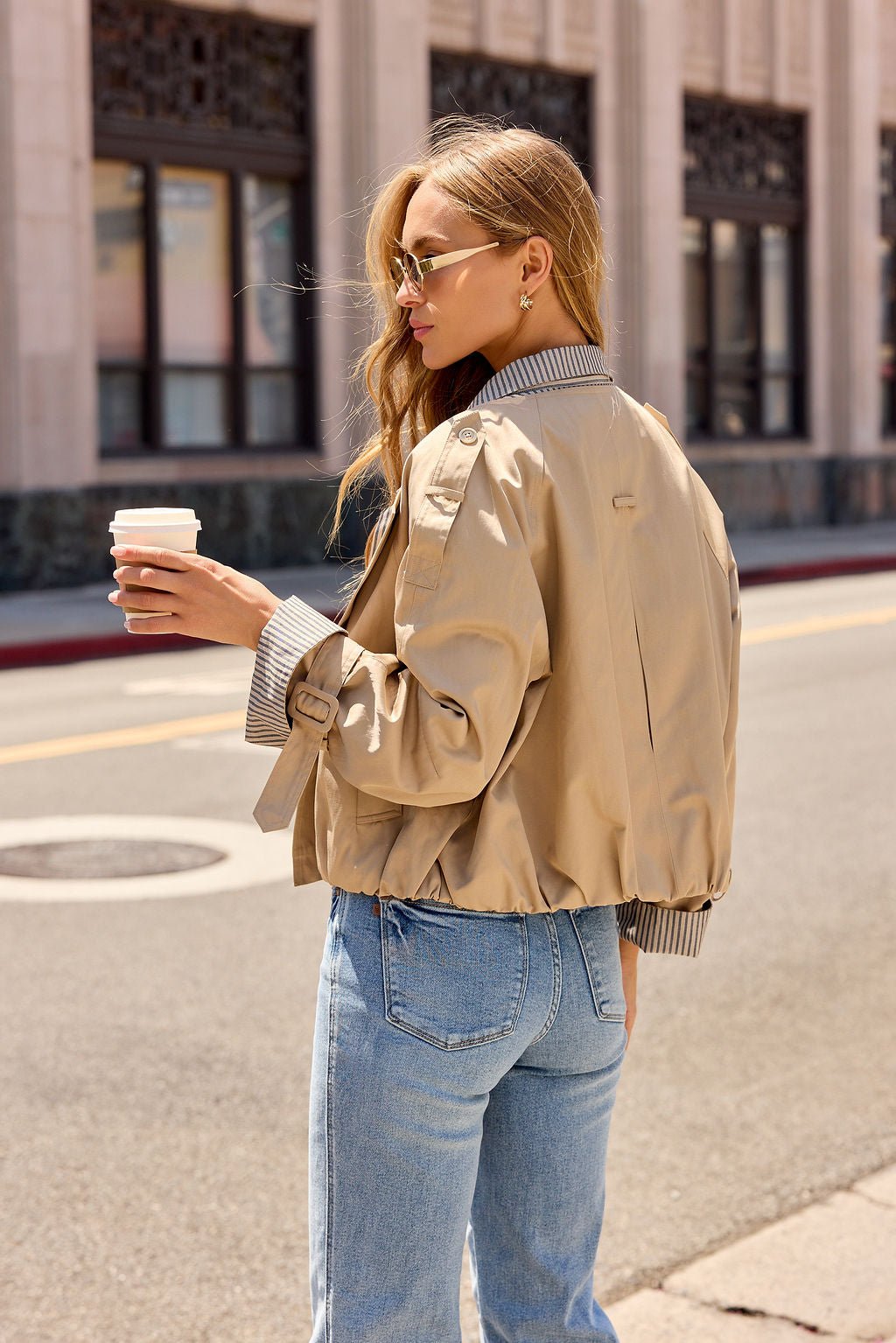 Woman in sunglasses, Contrast Striped Shirt Jacket - Camel, and jeans holds coffee on a city street in daylight.