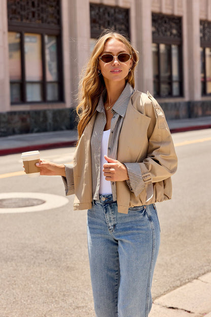 Woman in Contrast Striped Shirt Jacket - Camel, sunglasses, and jeans holds coffee on a city street with buildings behind her.