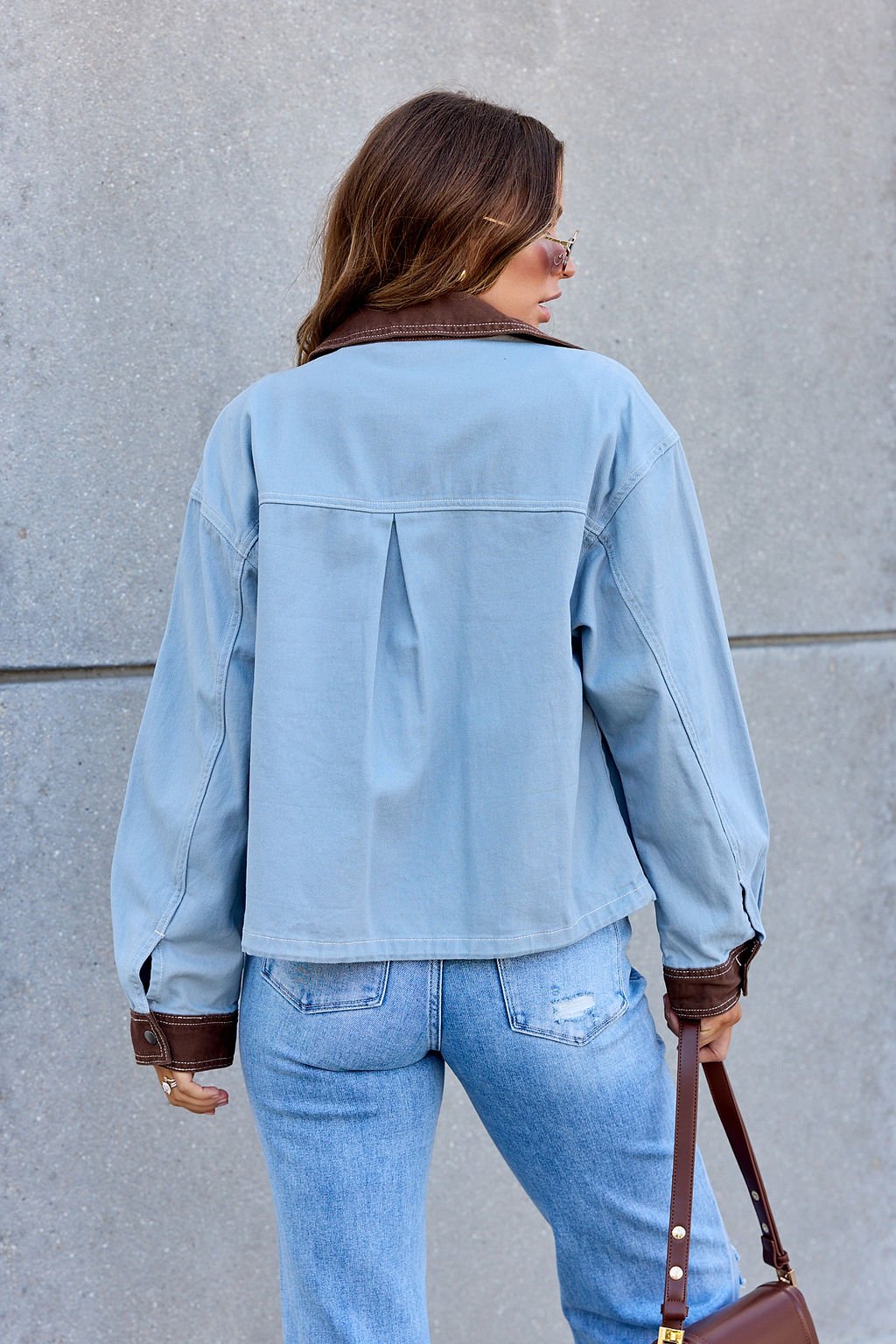Someone wears the Contrast Trim Jacket - Dusty Blue with jeans, facing a concrete wall and holding a brown purse.