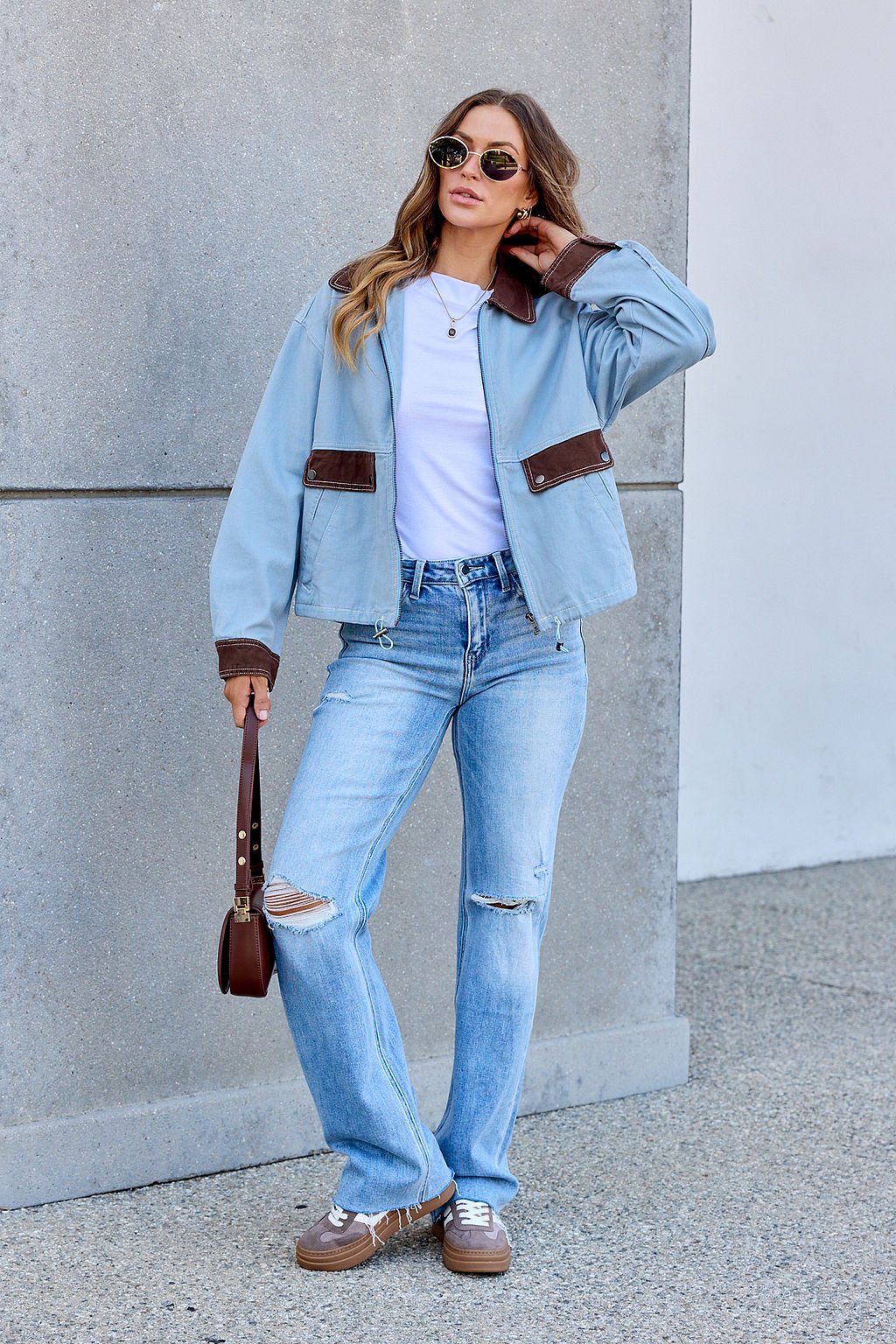 Woman in sunglasses wears a Contrast Trim Jacket - Dusty Blue, white shirt, ripped jeans, and holds a brown handbag by the wall.