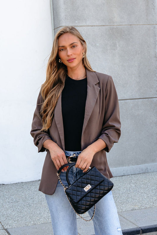 Woman in the Day To Night Blazer - Mocha with ruched sleeves, black top, jeans, and quilted purse, standing outdoors.