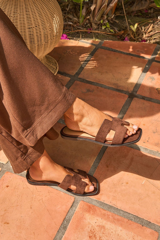 Wearing Desert Drift Sandals - Brown - FINAL SALE, seated on terracotta tiles and dressed in brown pants.