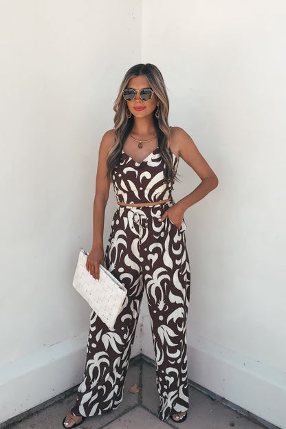 Woman in sunglasses wears the Eda Print Cami Tank and Pants Set in brown, holding a white clutch against a white wall.