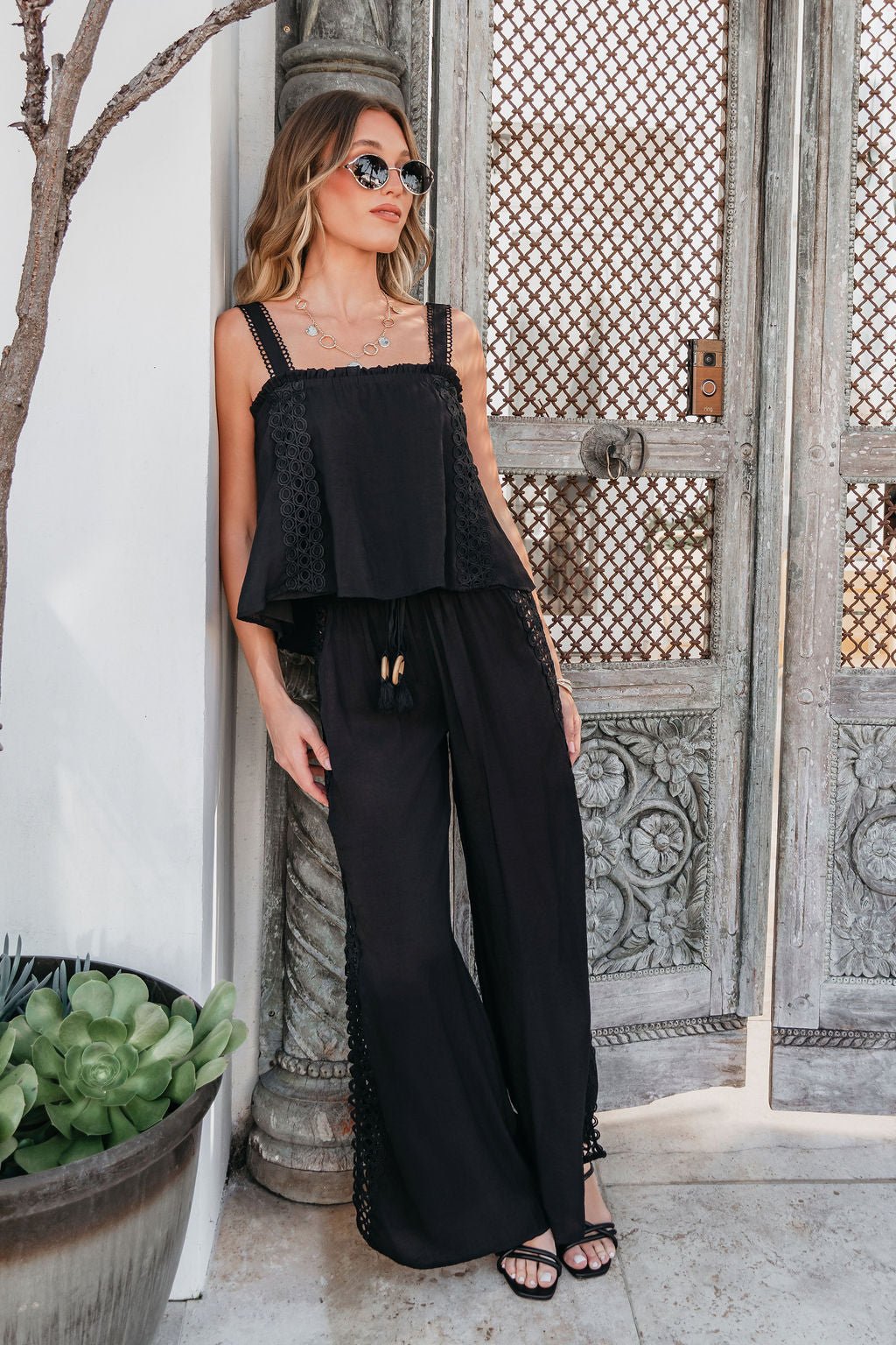 Woman in sunglasses wearing ELAN Black Lace Trim Wide Leg Pants, standing by ornate doors next to a potted plant.