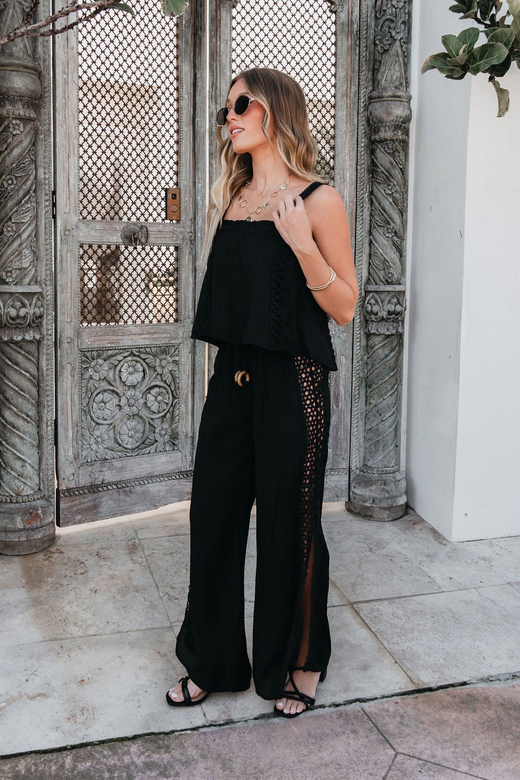 Woman in ELAN Black Lace Trim Wide Leg Pants and sandals stands outdoors by ornate doors, wearing sunglasses and minimal jewelry.