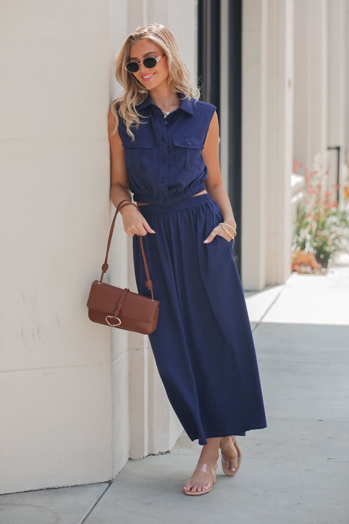 Woman wears the Elan Collared Button Down Top in navy, with sunglasses and sandals, smiling outdoors holding a brown handbag.