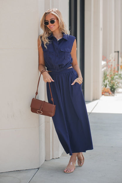 Woman wears the Elan Collared Button Down Top in navy, with sunglasses and sandals, smiling outdoors holding a brown handbag.