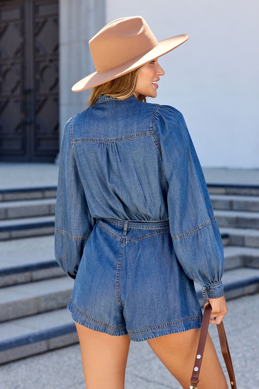 Woman in the Elan Denim Long Sleeve Belted Romper and tan hat stands outside, holding a purse by concrete steps, facing away.