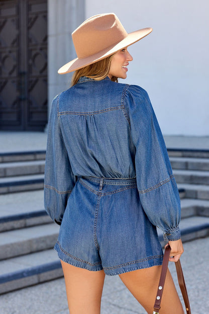 Woman in the Elan Denim Long Sleeve Belted Romper and tan hat stands outside, holding a purse by concrete steps, facing away.