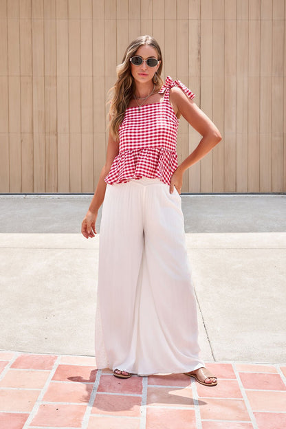 Woman in sunglasses wears a red gingham top and Elan Flowy Wide Leg Pants in ivory, standing on a tiled patio by a wooden wall.