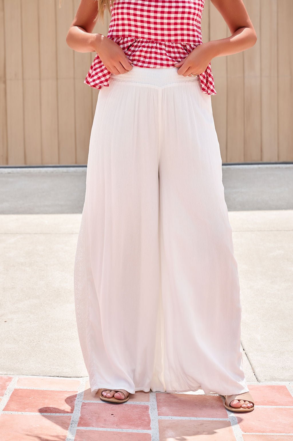 Person in a red gingham top and Elan Flowy Wide Leg Pants - Ivory adjusts the elastic waistband for a relaxed, flowy look outdoors.