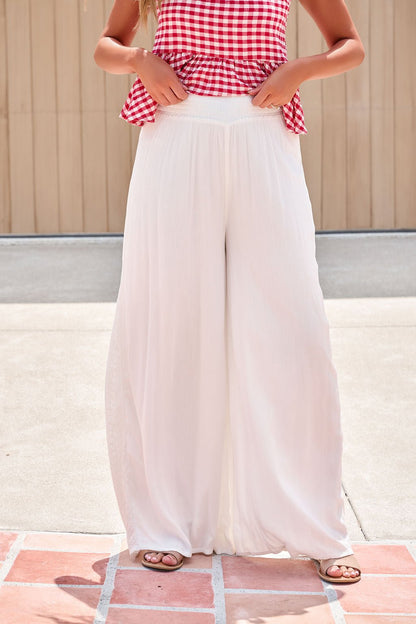 Person in a red gingham top and Elan Flowy Wide Leg Pants - Ivory adjusts the elastic waistband for a relaxed, flowy look outdoors.
