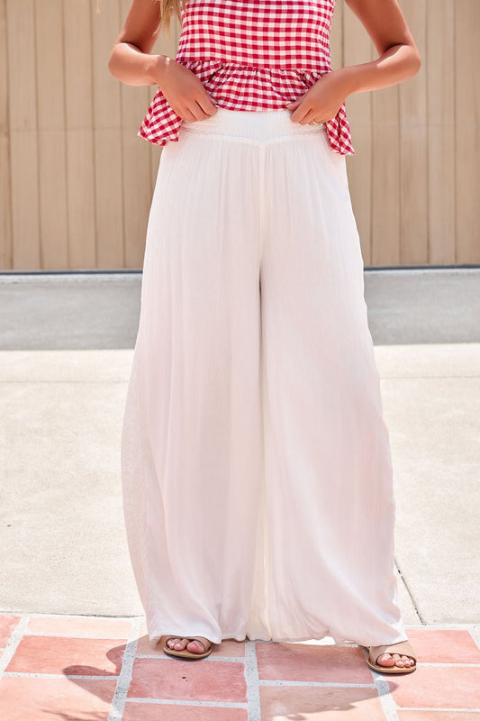 Person in a red gingham top and Elan Flowy Wide Leg Pants - Ivory adjusts the elastic waistband for a relaxed, flowy look outdoors.