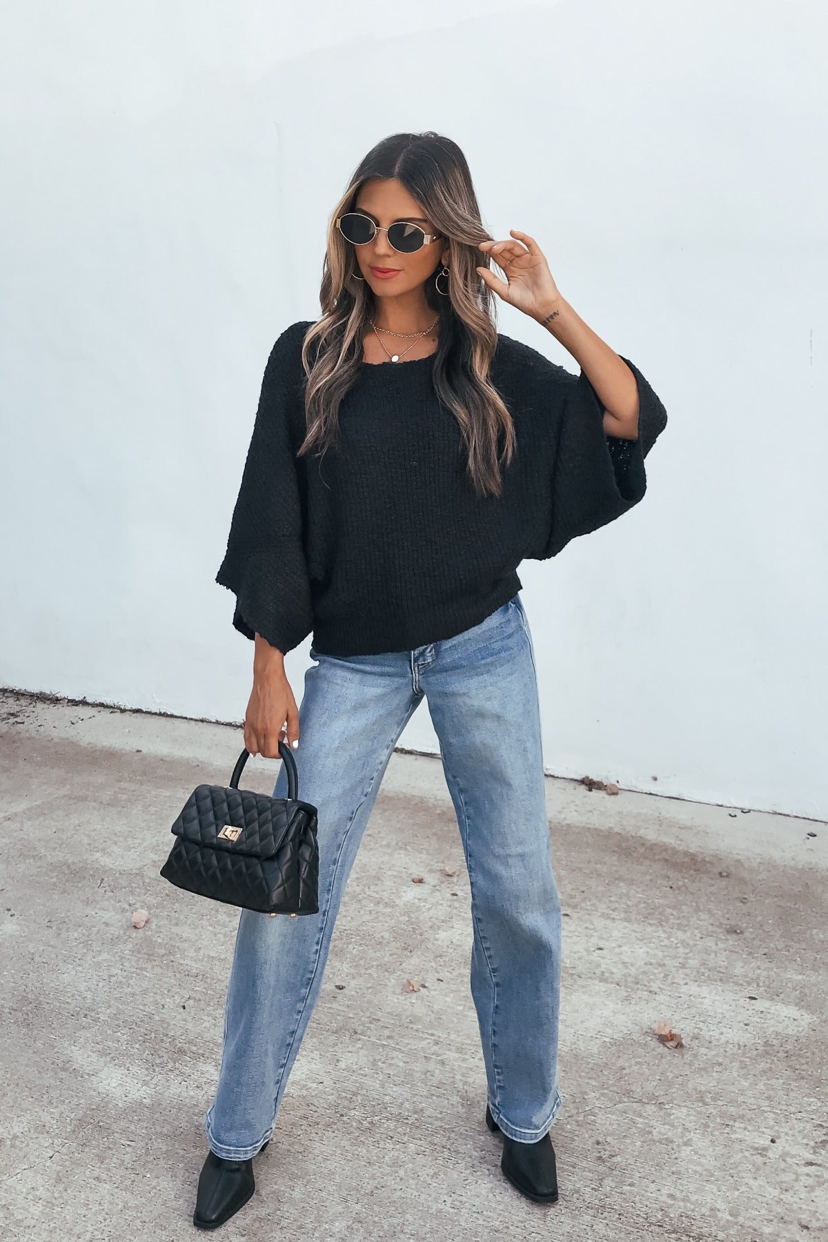 Woman in sunglasses and jeans poses confidently in an Elevated Ribbed Boatneck Sweater - Black, holding a quilted handbag by a white wall.