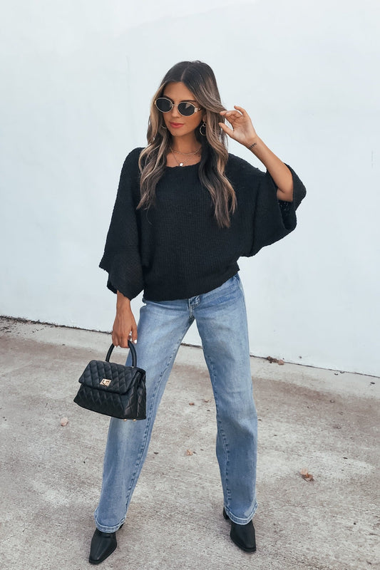 Woman in sunglasses and jeans poses confidently in an Elevated Ribbed Boatneck Sweater - Black, holding a quilted handbag by a white wall.