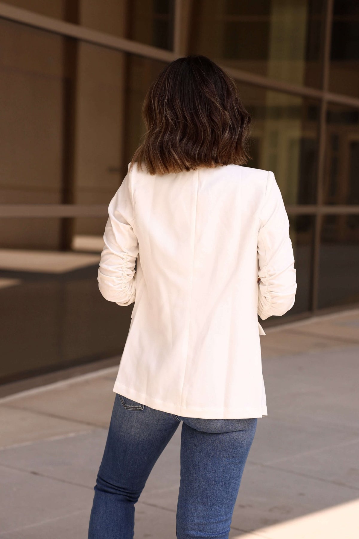 A person with wavy brown hair in a White Ruched Sleeve Blazer and blue jeans stands on a sidewalk near a glass building.