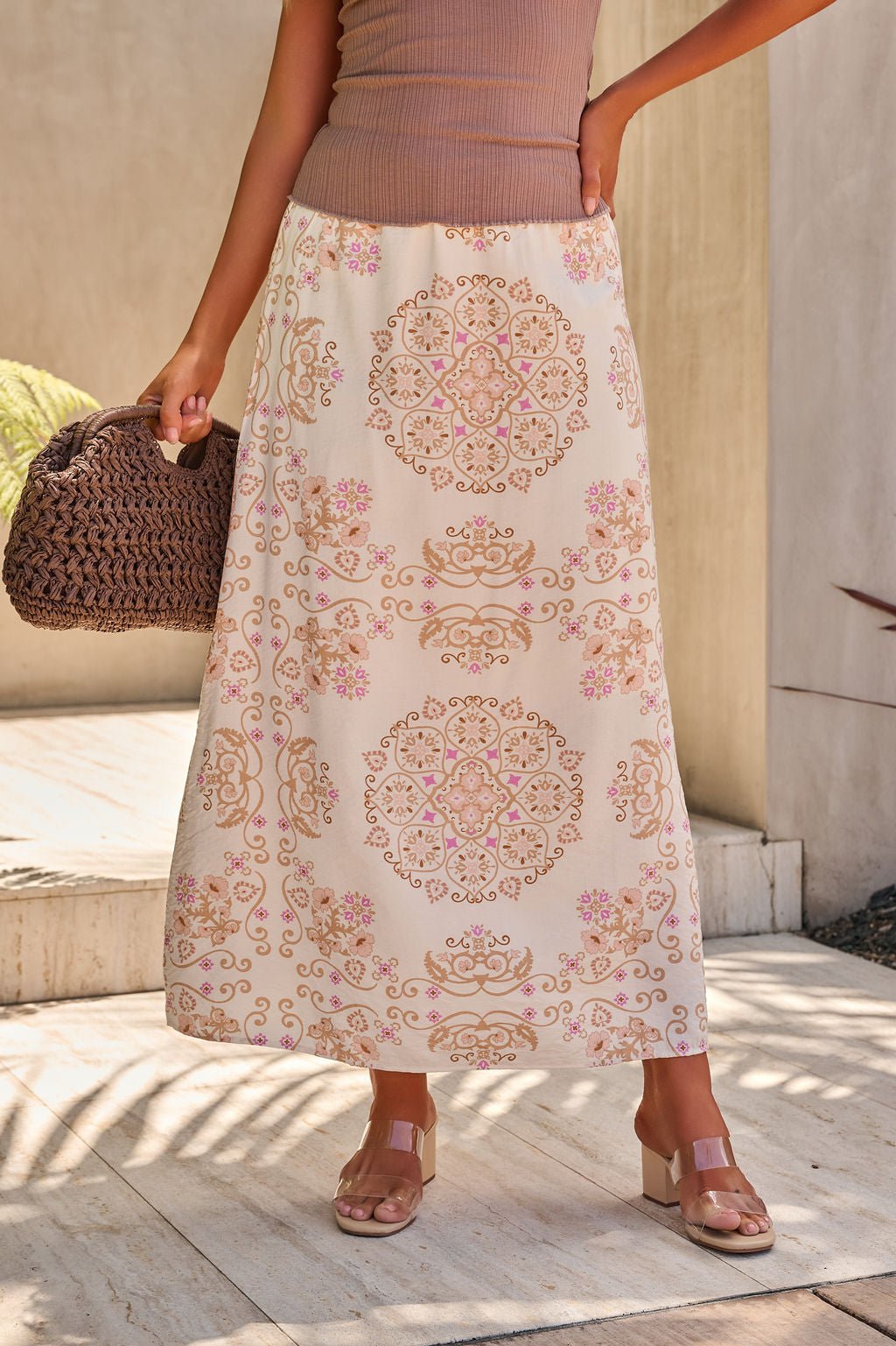 A woman wears the Evelyn Cream Print Maxi Skirt - FINAL SALE with sandals and a woven brown handbag.