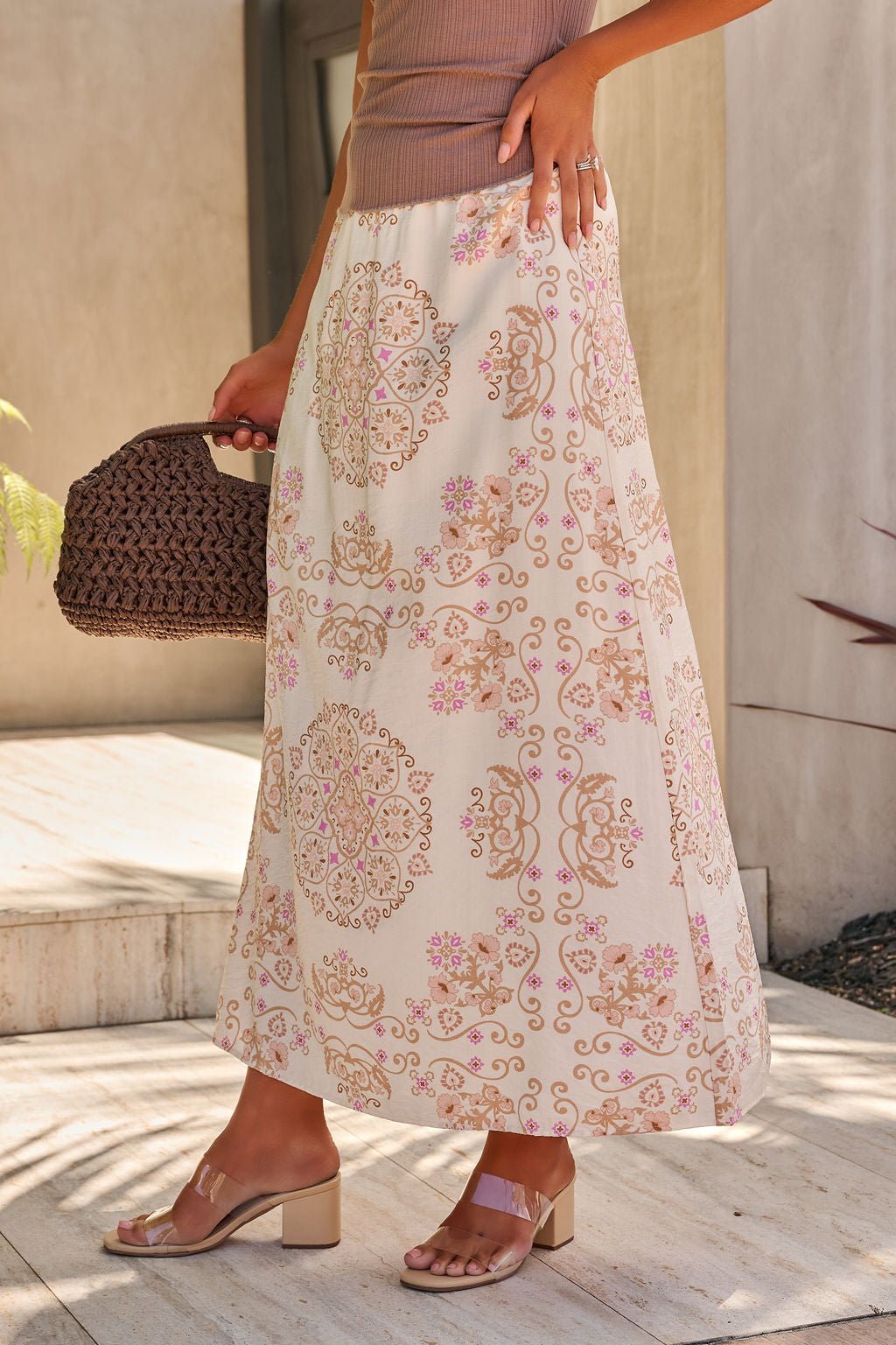 Woman wears the Evelyn Cream Print Maxi Skirt - FINAL SALE with an elastic waistband, sandals, and a brown woven handbag outdoors.