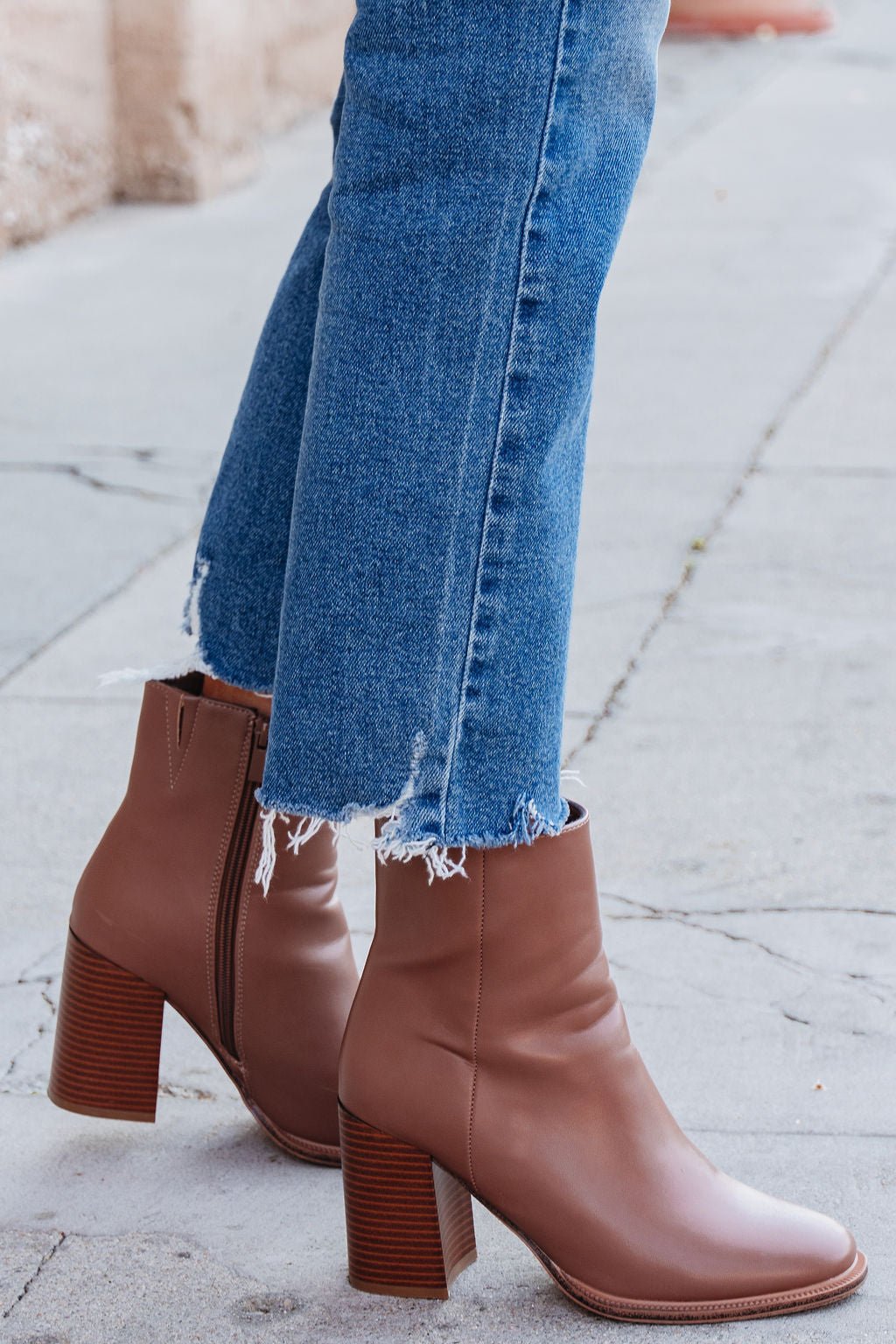 Person in frayed hem blue jeans and the Evelyn Taupe Leather Heeled Ankle Booties stands on a sidewalk.