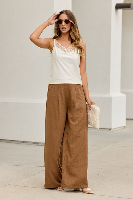Woman in sunglasses wears the Evening Lace Satin Tank Top in Champagne, wide brown pants, and holds a woven clutch on a city sidewalk.