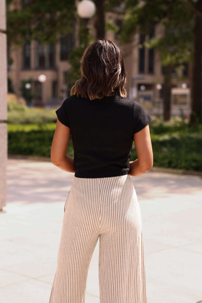 A woman wears the Everyday Essential Black Tee-FINAL SALE with striped pants outdoors. The 100% cotton top ensures a comfy fit.