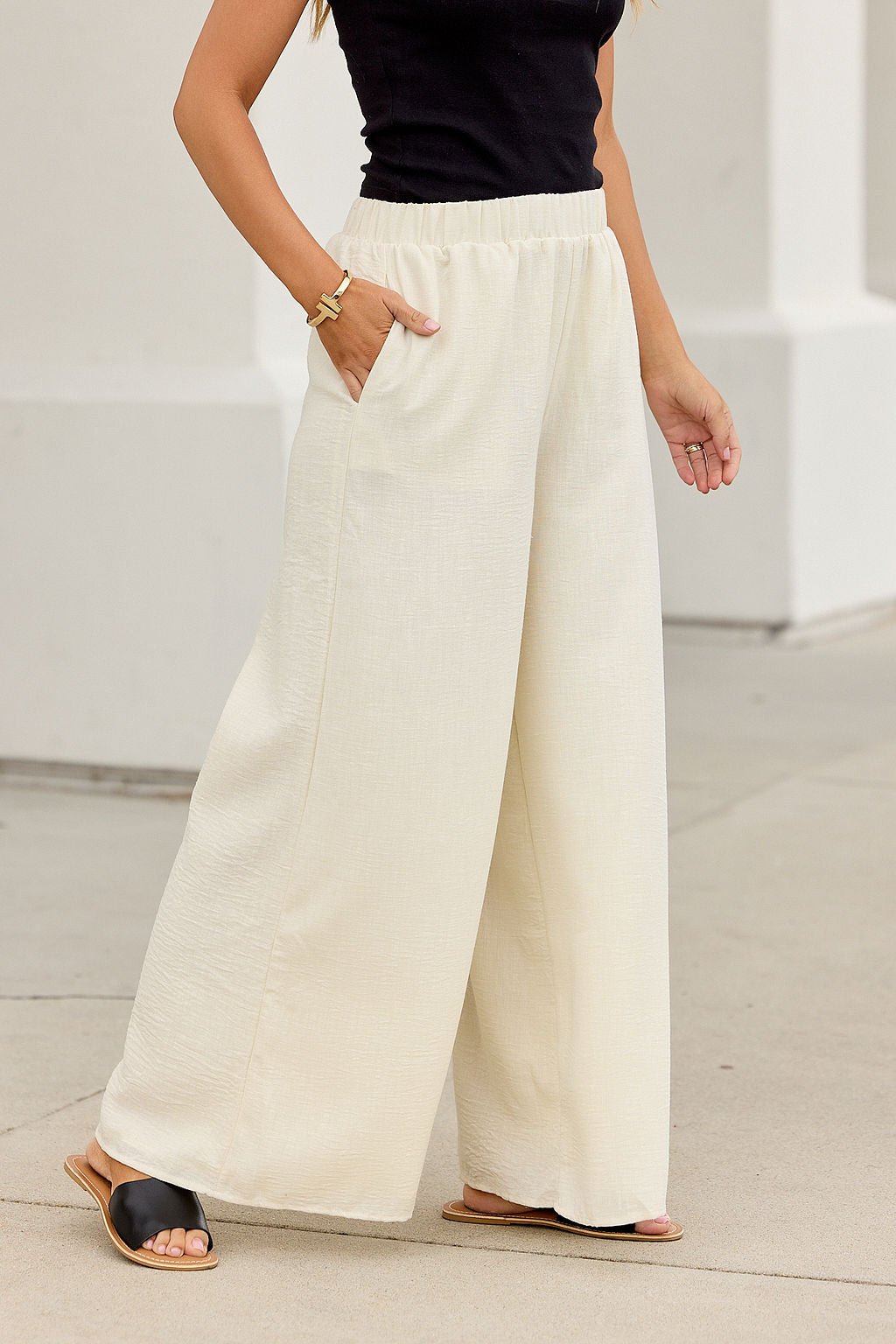 Woman in First Class Wide Leg Pants - Natural poses stylishly on the sidewalk, paired with a black top and gold bracelet.