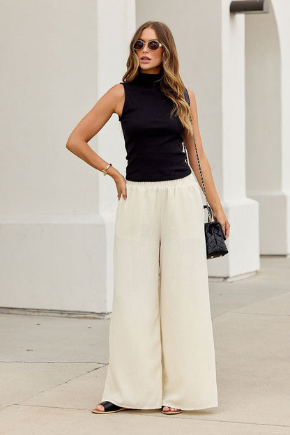 Woman in sunglasses and black top wears First Class Wide Leg Pants - Natural, holding a purse on a city sidewalk.