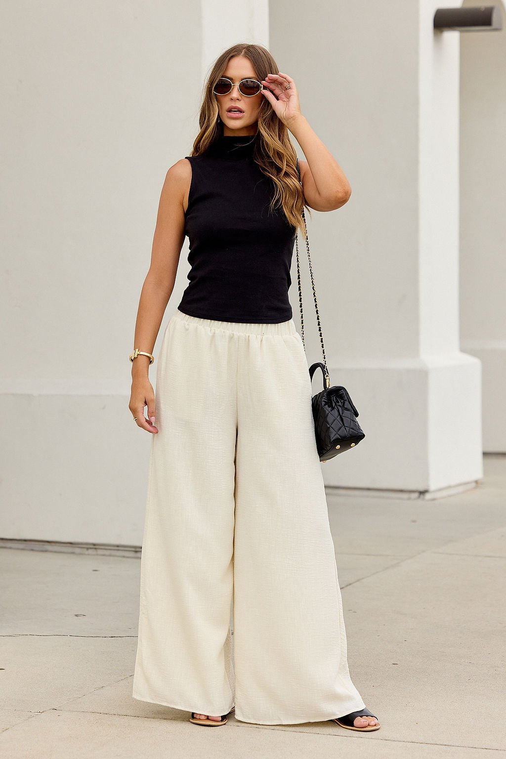 Woman in sunglasses, sleeveless black top, First Class Wide Leg Pants - Natural, black sandals, and purse on a city sidewalk.