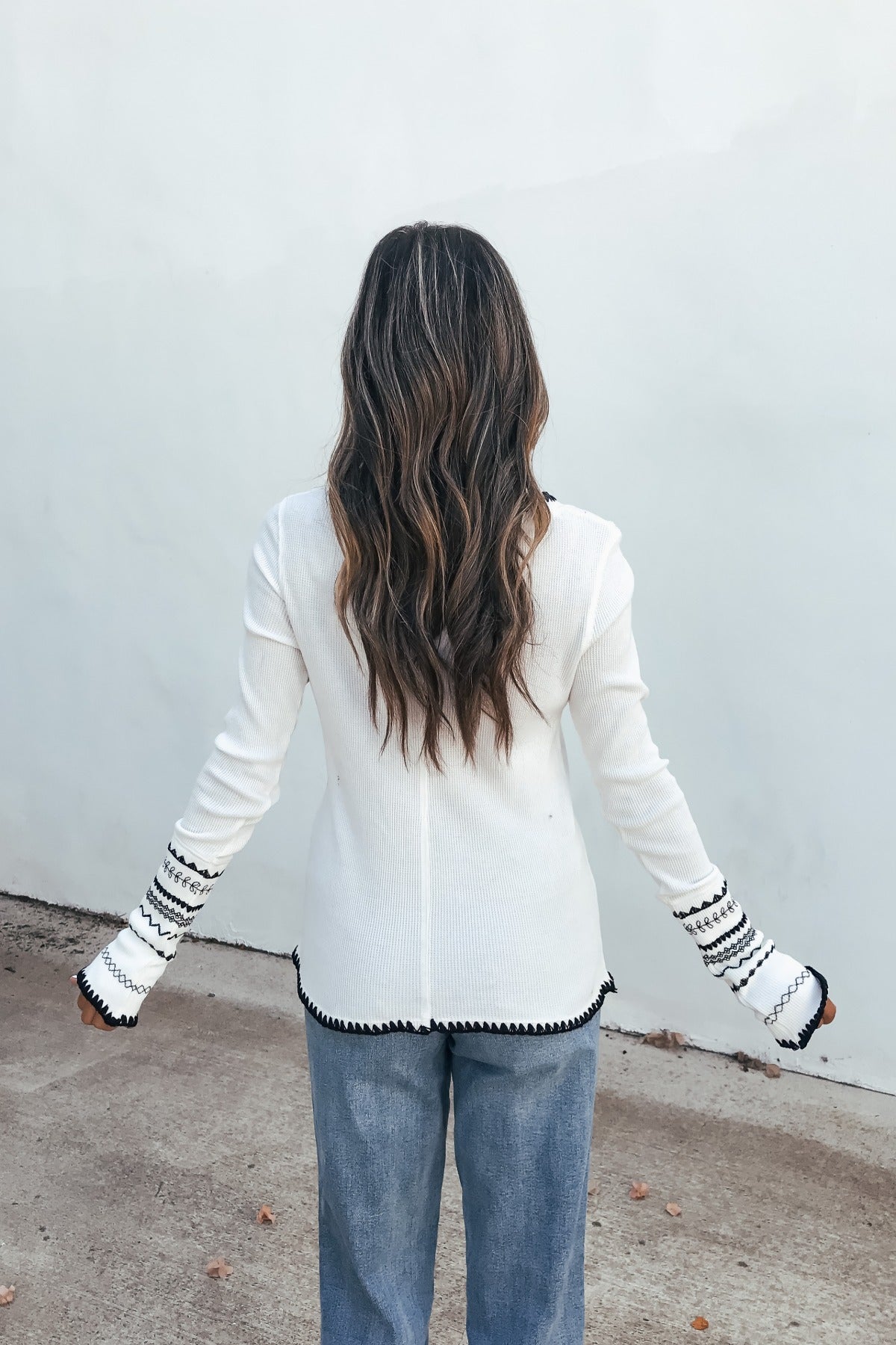 Person with long wavy hair in a white Free People Aspen Thermal Top and blue jeans, standing outdoors facing away.