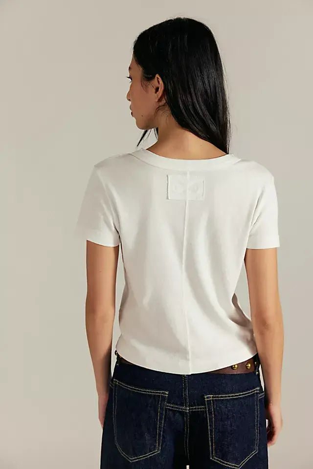 Someone in a fitted ivory Free People Atlas Crew Tee and dark jeans stands with their back to the camera, long dark hair flowing.