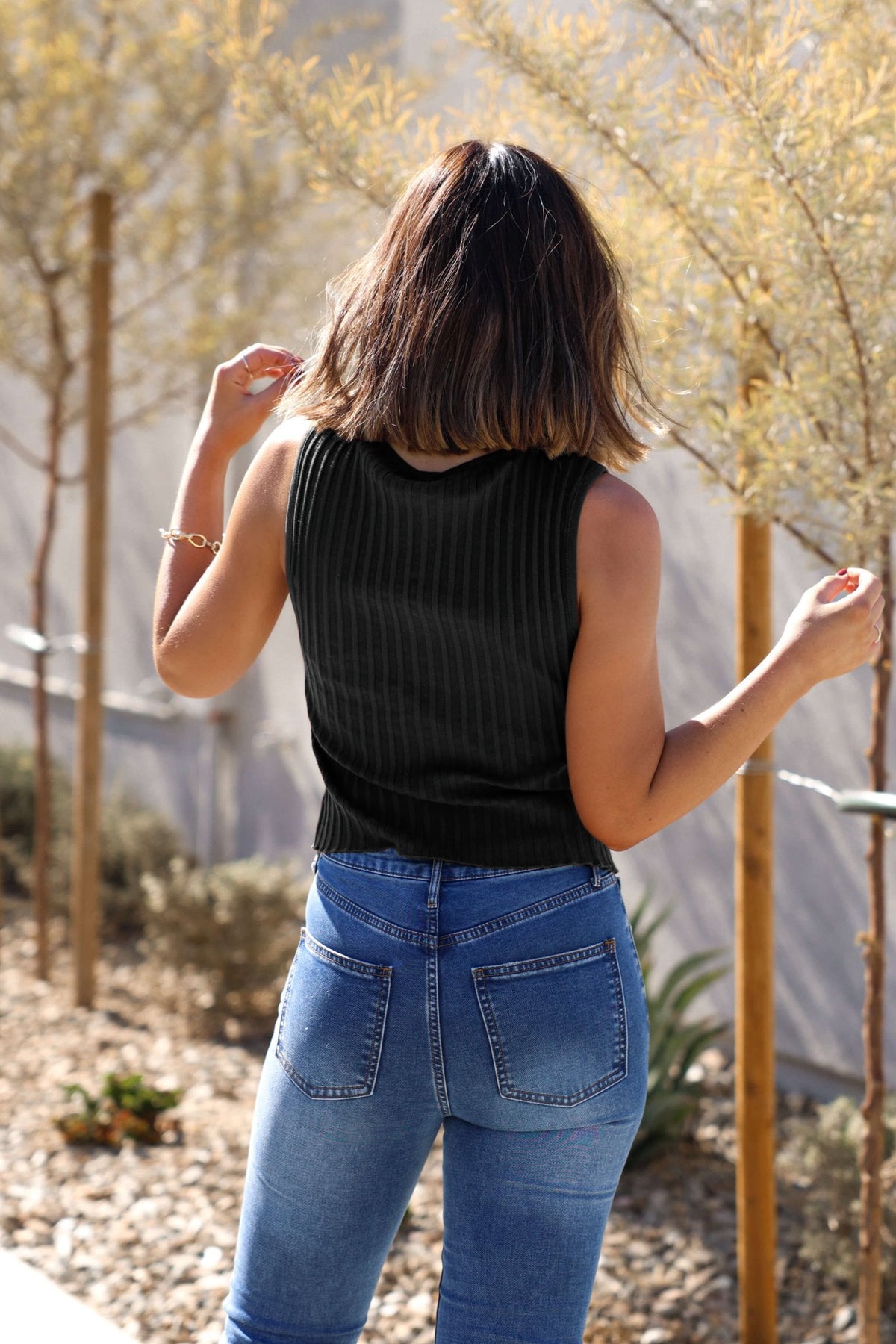 Woman with shoulder-length brown hair wears a slim fit Free People Black Tilly Vest Top and blue jeans, outdoors among small trees.