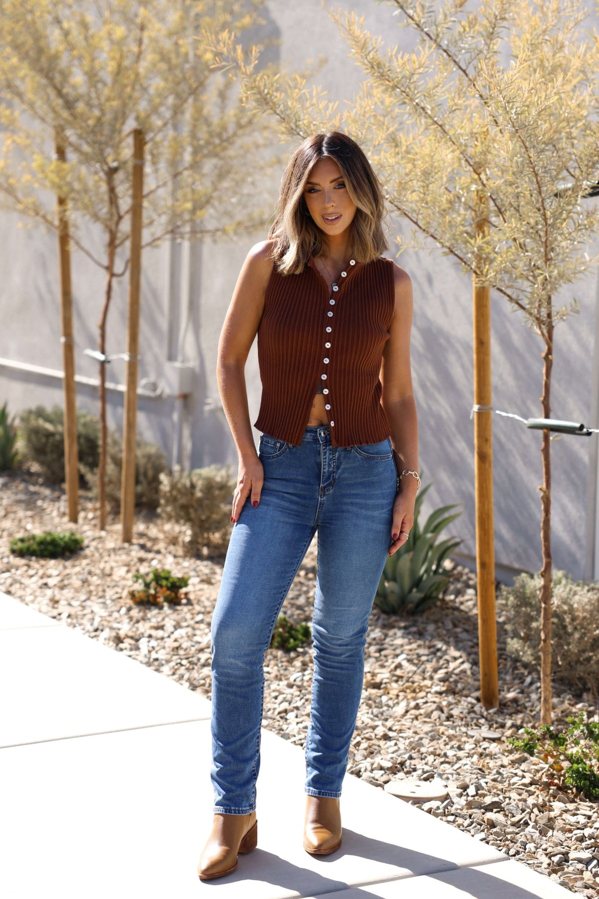 Woman wears a Free People Brown Tilly Vest Top, blue jeans, and boots on a sidewalk lined with small trees and plants.
