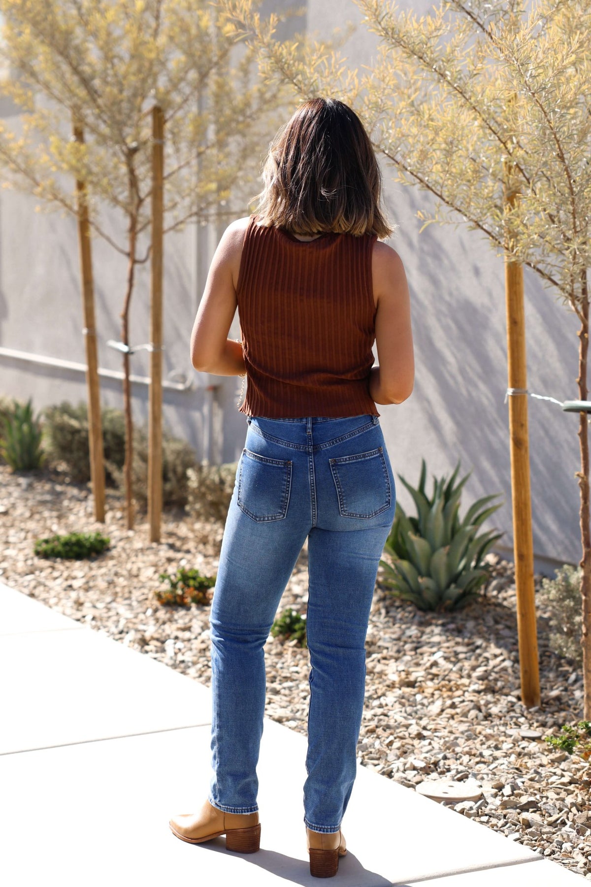 Model wears the Free People Brown Tilly Vest Top on a sidewalk, paired with blue jeans, facing away by plants.